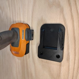 May include: An orange and black cordless drill with the word "BRUSHLESS" printed on the side is shown next to a black mounting bracket. The drill is partially inserted into the bracket, suggesting a storage or mounting solution.