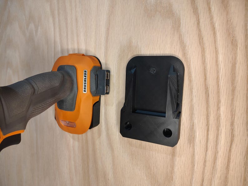 May include: An orange and black cordless drill with the word "BRUSHLESS" printed on the side is shown next to a black mounting bracket. The drill is partially inserted into the bracket, suggesting a storage or mounting solution.