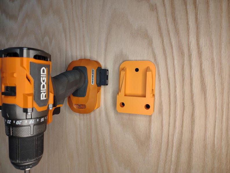 May include: An orange and black Ridgid brand drill with a matching orange battery and wall mount. The drill is partially visible, with the battery and mount attached to a wooden surface.