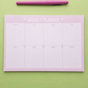 May include: A pink weekly planner with a floral design. The planner has spaces for each day of the week, Monday through Sunday, and a notes section.