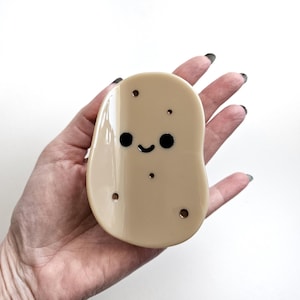 Può includere: Un oggetto a forma di patata color beige, con un volto sorridente e piccoli puntini marrone scuro. L'oggetto ha una finitura lucida. La patata ha un design a cartone animato ed è tenuta in mano su sfondo bianco.