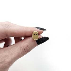 Puede incluir: Un pin de esmalte dorado en forma de patata con una cara sonriente y pequeños puntos blancos. El pin se sostiene entre los dedos sobre un fondo blanco. El pin es un accesorio lindo y caprichoso.