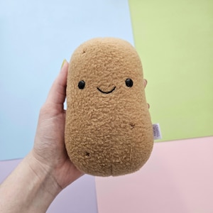Puede incluir: Un peluche de patata de color marrón claro con una cara sonriente y pequeños ojos negros. La patata tiene una superficie texturizada. Una pequeña etiqueta está adherida al lateral.