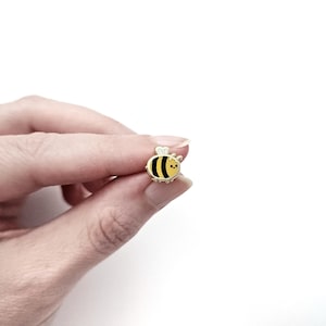 Puede incluir: Un pequeño pin de esmalte con forma de abeja, con un cuerpo a rayas negras y amarillas, una cara sonriente y detalles dorados. El pin se sostiene entre los dedos sobre un fondo blanco.
