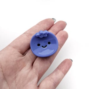May include: A small, round, light purple blueberry-shaped pin with a smiling face and a small flower on top. The pin is smooth and shiny, with black dots for eyes and a curved black line for a mouth. The pin is held in a person's hand.
