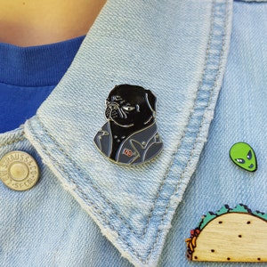 Puede incluir: Un pin de un pug negro con una chaqueta de cuero gris con la palabra "NO" en ella. El pug lleva un collar de cadena de plata. El pin está en una chaqueta vaquera.