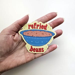 May include: A die-cut sticker featuring a blue bowl filled with refried beans. The bowl has a smiling face and heart details. The words "refried beans" are in red, with yellow star accents.