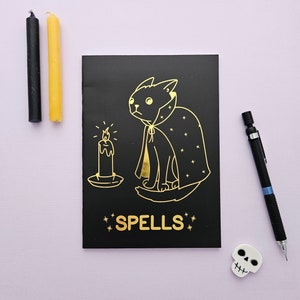 May include: A black notebook with a gold illustration of a cat wearing a cape and a candle. The notebook says "SPELLS" in gold lettering.