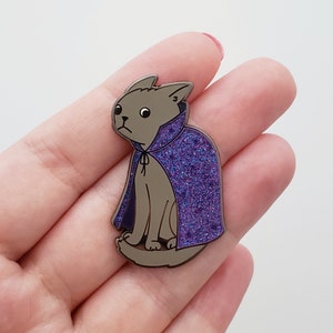May include: A silver enamel pin of a gray cat wearing a purple glitter cape. The cat has a sad expression on its face.