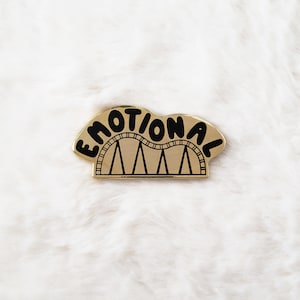 May include: Gold-colored pin with the word "EMOTIONAL" written in black letters in a wavy font. The word is on top of a black roller coaster track with three gold supports.