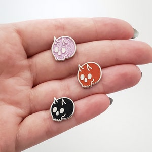 Puede incluir: Tres pines de esmalte con forma de calaveras de dibujos animados con cuernos. Las calaveras son rosas, naranjas y negras.