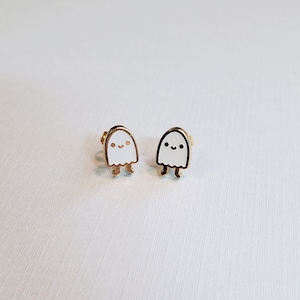 Puede incluir: Un par de pendientes de botón de tono dorado con un diseño de fantasma de esmalte blanco con una cara sonriente.