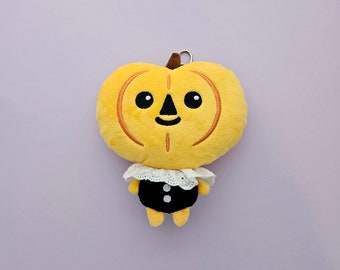 Puede incluir: Un juguete de peluche con forma de calabaza con una cara en blanco y negro y un cuello de volantes blanco y negro. El juguete lleva un vestido negro con dos botones blancos.