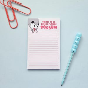 Peut inclure: Un bloc-notes rose avec des lignes horizontales et le texte "THINGS TO DO BEFORE PLAYING POSSUM" et une illustration d'opossum. Un stylo bleu clair avec des visages souriants et deux trombones rouges sont également dans l'image.