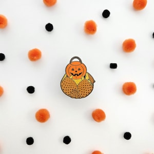 May include: A brown and orange enamel pin of a pumpkin in a brown and orange cornucopia. The pin is on a white background with orange and black polka dots.
