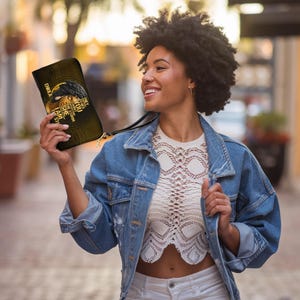 Puede incluir: Un bolso de mano negro y dorado con una correa de mu&ntilde;eca. El bolso presenta una ilustraci&oacute;n del rostro de una mujer y el texto "EMBOLA, THEY TRIED TO BURY YOU, THEY DIDN'T KNOW YOU WERE A SEED".