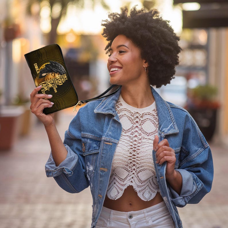 Puede incluir: Un bolso de mano negro y dorado con una correa de mu&ntilde;eca. El bolso presenta una ilustraci&oacute;n del rostro de una mujer y el texto "EMBOLA, THEY TRIED TO BURY YOU, THEY DIDN'T KNOW YOU WERE A SEED".