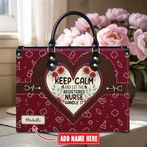Könnte beinhalten: Burgunderfarbene Tragetasche mit schwarzem Griff und goldfarbener Hardware. Die Tasche hat ein herzförmiges Design mit dem Text "KEEP CALM AND LET THE REGISTERED NURSE HANDLE IT". Außerdem gibt es medizinische Symbole und Platz für einen Namen.