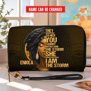 Puede incluir: Cartera negra y dorada con cierre de cremallera y correa de mu&ntilde;eca. La cartera presenta un retrato de una mujer con el texto "THEY WHISPERED TO HER YOU CANNOT WITHSTAND THE STORM SHE WHISPERED BACK I AM THE STORM" y el nombre "ENIOLA".