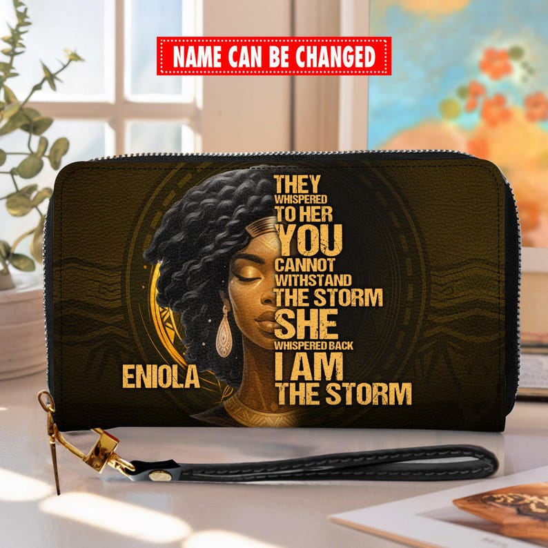 Puede incluir: Cartera negra y dorada con cierre de cremallera y correa de mu&ntilde;eca. La cartera presenta un retrato de una mujer con el texto "THEY WHISPERED TO HER YOU CANNOT WITHSTAND THE STORM SHE WHISPERED BACK I AM THE STORM" y el nombre "ENIOLA".