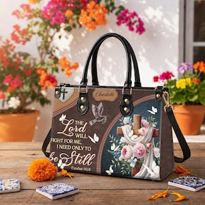 Personalized Christian tote bag with praying angel artwork, soft pink and white flowers, cross accents, and the inspirational Bible quote “The Lord will fight for you, you need only be still.” Designed with black handles and a structured shape, this religious handbag makes a thoughtful faith gift for women, mom, grandma, wife, or daughter for birthdays, Christmas, Mother’s Day, or church celebrations.