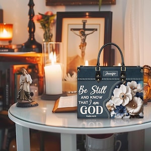 Puede incluir: Una bolsa de mano azul oscuro con un dise&ntilde;o floral y el texto "Be Still and Know That I am God" en blanco. La bolsa tiene un asa negra y una etiqueta de nombre que dice "Jennifer".