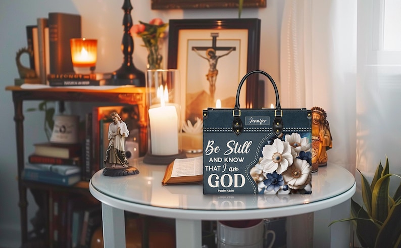 Puede incluir: Una bolsa de mano azul oscuro con un dise&ntilde;o floral y el texto "Be Still and Know That I am God" en blanco. La bolsa tiene un asa negra y una etiqueta de nombre que dice "Jennifer".