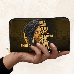 Puede incluir: Cartera negra y dorada con cierre de cremallera. La cartera presenta un retrato de una mujer con el texto "They whispered to her, you cannot withstand the storm. She whispered back, I am the storm." El nombre "Eniola" tambi&eacute;n est&aacute; impreso.