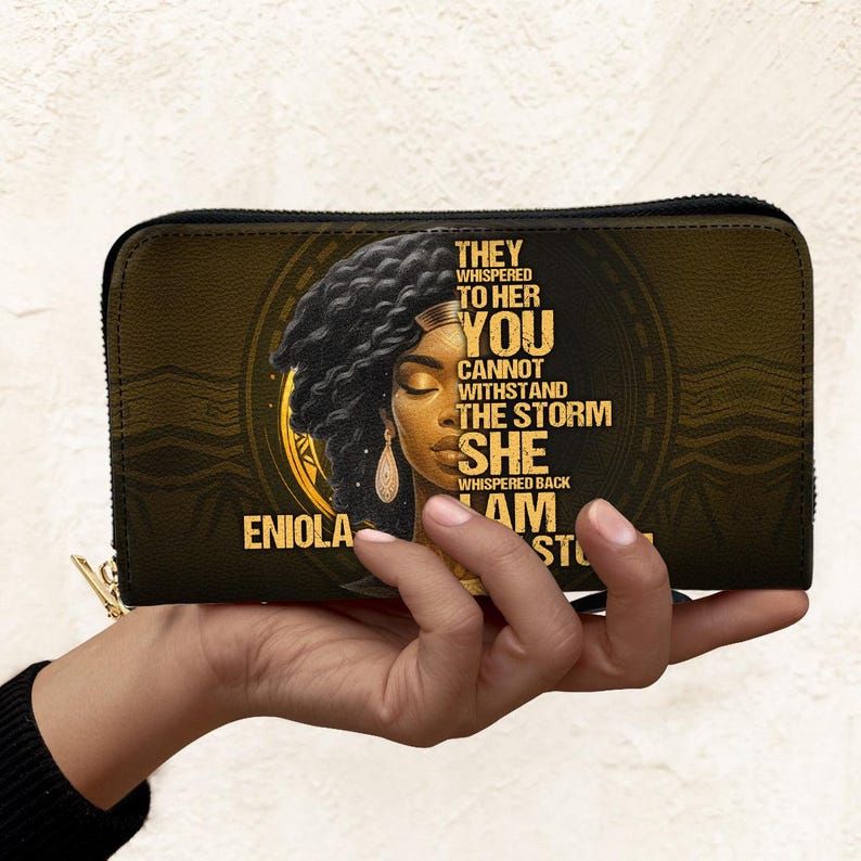 Puede incluir: Cartera negra y dorada con cierre de cremallera. La cartera presenta un retrato de una mujer con el texto "They whispered to her, you cannot withstand the storm. She whispered back, I am the storm." El nombre "Eniola" tambi&eacute;n est&aacute; impreso.