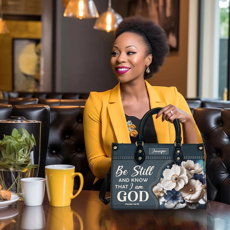 Puede incluir: Un bolso negro con un dise&ntilde;o floral y el texto "Be Still and Know That I am God, Psalm 46:10". El bolso tiene una cremallera dorada y un asa dorada. El bolso est&aacute; sobre una mesa con una taza de caf&eacute; y un plato de comida.