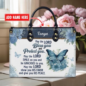 Pode incluir: Bolsa branca com alça preta e parte superior semelhante a jeans. A bolsa apresenta desenhos florais azuis e uma borboleta. O texto na bolsa diz "May the LORD Bless you and Protect you." O nome "Tonya" também é exibido.
