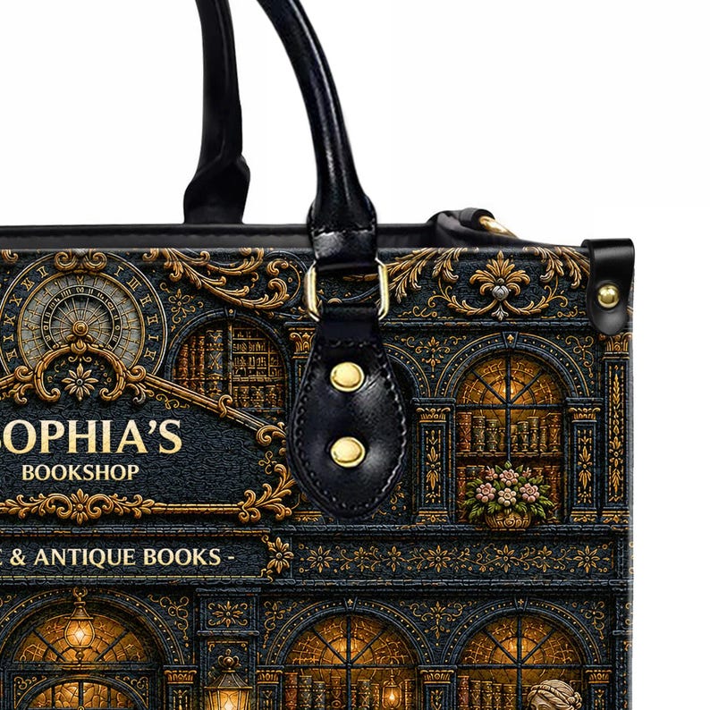 Puede incluir: Bolso tote negro con una ilustraci&oacute;n detallada de una fachada de librer&iacute;a, con ventanas arqueadas, detalles dorados ornamentados y el texto "SOPHIA'S BOOKSHOP & ANTIQUE BOOKS". El bolso tiene asas negras y herrajes dorados.