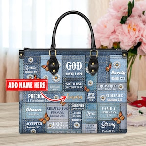 May include: A denim handbag with a patchwork design featuring inspirational quotes and bible verses. The bag has a black handle and a gold zipper. The bag is decorated with butterflies and daisies.