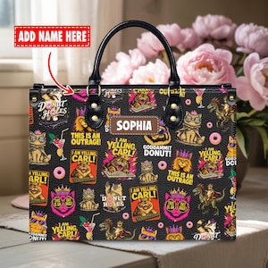 Könnte beinhalten: Eine schwarze Handtasche mit einem wiederkehrenden Grafikdruck von Katzen, Dinosauriern und Donuts sowie Texten wie "DONUT HOLES", "I AM YELLING CARL", "THIS IS AN OUTRAGE" und "GODDAMMIT DONUT!". Ein zentraler Aufnäher zeigt den Namen "SOPHIA".