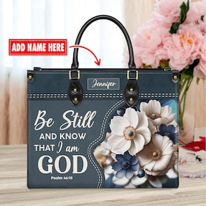 May include: A blue tote bag with a floral design and the text "Be Still and Know That I am GOD Psalm 46:10". The bag has a black handle and a gold nameplate that says "Jennifer".