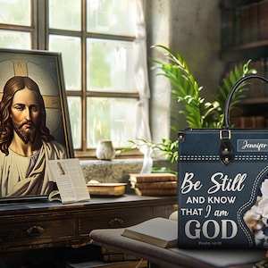 Puede incluir: Una bolsa de mano azul con un dise&ntilde;o floral y el texto "Be Still and Know That I am GOD" con una referencia b&iacute;blica. La bolsa tiene un asa negra y una etiqueta de nombre personalizada.