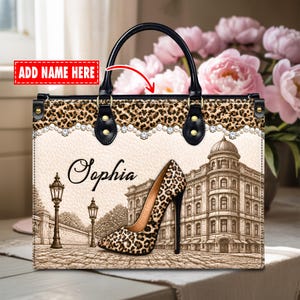May include: A rectangular handbag with black handles and a leopard print top border accented by a row of white pearls. The front features a beige background with a printed illustration of a leopard print high heel, street lamps, and a building.