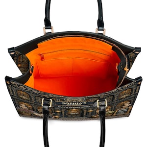 Puede incluir: Bolso tote negro con un dise&ntilde;o detallado de librer&iacute;a. El bolso tiene asas negras y herrajes dorados. El interior es de color naranja brillante con bolsillos y una cremallera. El texto "SOPHIA'S BOOKSHOP RARE & ANTIQUE BOOKS" es visible.
