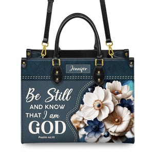 Puede incluir: Un bolso de mano de mezclilla azul con asa negra y detalles dorados. El bolso presenta un dise&ntilde;o floral con flores blancas y azules. El texto "Be Still and Know That I am God" est&aacute; impreso en la parte delantera del bolso, junto con el vers&iacute;culo "Salmo 46:10". El nombre "Jennifer" est&aacute; impreso en una placa de identificaci&oacute;n en la parte delantera del bolso.