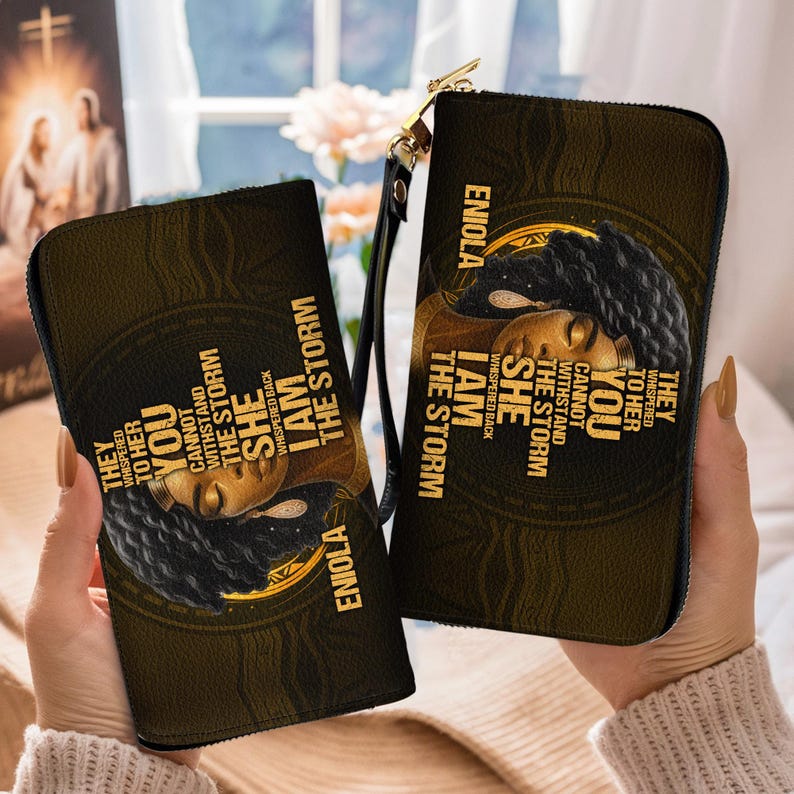 Puede incluir: Dos carteras negras y doradas con cremallera con un retrato de mujer y el texto "I AM THE STORM". Las carteras tienen una correa de mu&ntilde;eca y una cremallera dorada. El texto "ENIOLA" tambi&eacute;n es visible.