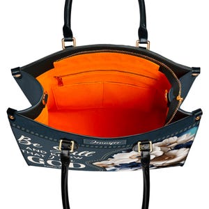 Puede incluir: Un bolso tote azul con estampado floral y el texto "Be Still and Know that I am God" en blanco. El bolso tiene cierre de cremallera y forro naranja.