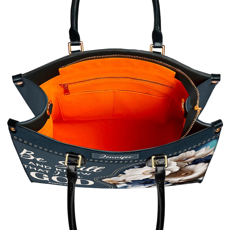 Puede incluir: Un bolso tote azul con estampado floral y el texto "Be Still and Know that I am God" en blanco. El bolso tiene cierre de cremallera y forro naranja.
