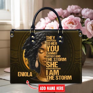 Pode incluir: Bolsa tote de couro preta com um design dourado e marrom apresentando um retrato de mulher e o texto "THEY WHISPERED TO HER YOU CANNOT WITHSTAND THE STORM SHE WHISPERED BACK I AM THE STORM". A bolsa tem uma alça preta e ferragens douradas.