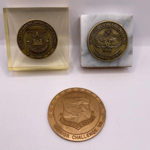 May include: Three bronze military challenge coins. One coin is encased in a clear acrylic block, another is set on a marble tile, and the third is displayed on its own. The coins feature text and emblems.