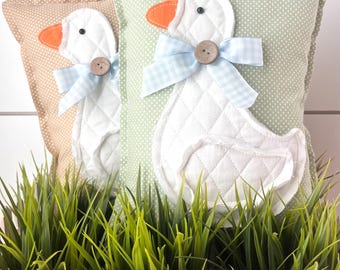 May include: Two decorative pillows featuring a white quilted goose design with orange beaks, black eyes, and light blue gingham bows. One pillow is tan with white polka dots, the other is light green with white polka dots. The pillows are displayed in front of a white picket fence and green grass.