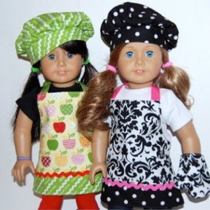 May include: Two dolls wearing chef hats and aprons. One doll is wearing a green and yellow apple-print apron and a green chef hat. The other doll is wearing a black and white damask-print apron and a black and white polka dot chef hat.