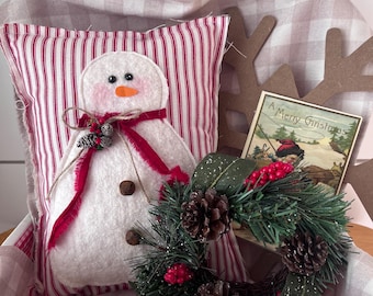 May include: A festive Christmas display featuring a snowman pillow with a red striped background, a small wreath with pinecones and berries, and a vintage Christmas card. The arrangement is set in a basket with a checkered fabric.
