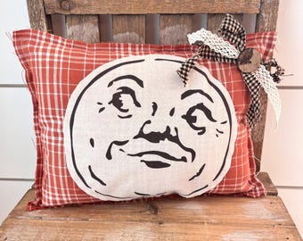 May include: A rectangular throw pillow featuring a red and white checkered pattern with a large, circular, off-white patch printed with a black graphic of a moon face. The pillow is accented with a black and white gingham ribbon and lace bow.