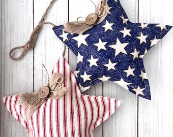 May include: Two star-shaped patriotic ornaments. One is navy blue with white stars, the other is red and white striped. Each has a jute bow and button detail, with a jute loop for hanging. The background is a white wooden surface.