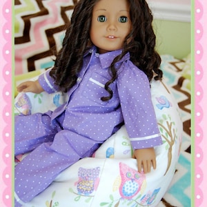 May include: A doll with long, dark curly hair wearing purple polka dot pajamas with white trim. The doll is seated on a white and cream-colored beanbag chair decorated with colorful owl and tree designs.